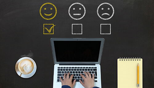 Person typing on a laptop with customer feedback questions represented by smiley, neutral, and sad face icons, symbolizing different levels of customer satisfaction