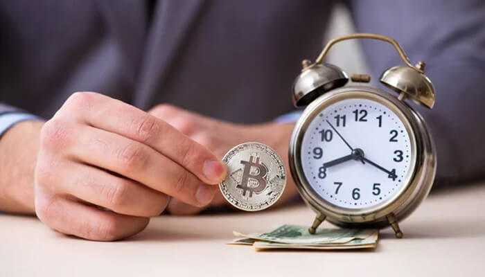 Person holding a bitcoin coin next to an analog clock and cash to represent the concept of the best time to buy cryptocurrency.