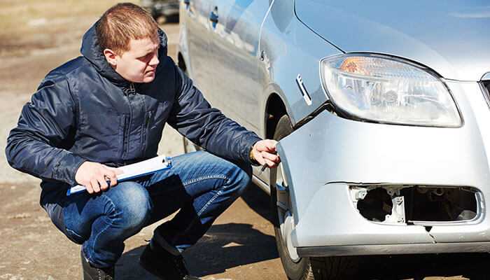 A man inspecting damage to the front bumper and side of a silver car after a car accident, holding a clipboard while crouching down, representing insurance work in a car accident