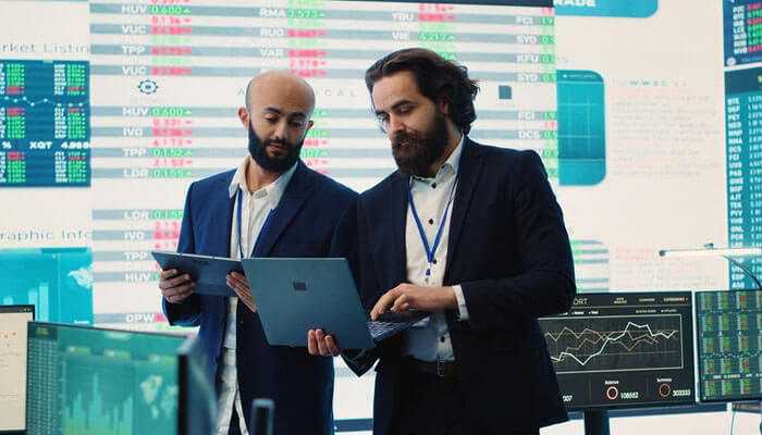 Business professionals reviewing financial data on a laptop discuss decision making in hedge funds with the stock charts in the background.