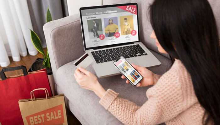 Customer shopping online with woocommerce plugins on a laptop and smartphone, browsing a fashion stores sale section with shopping bags nearby, representing an enhanced ecommerce experience