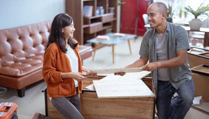 Working together, two colleagues put together custom made office furniture in a cozy, modern living room setting.