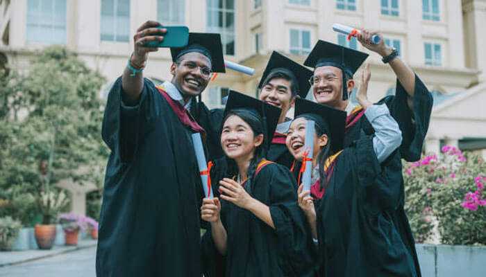 Diverse graduates in caps, gowns, and graduation sashes taking a selfie, celebrating with diplomas.