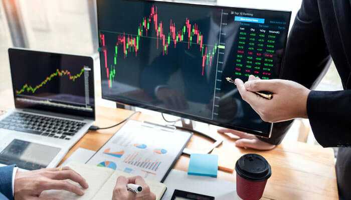 The image shows two people analyzing stock charts and financial data on computer screens, likely discussing investment strategies or stock market trends