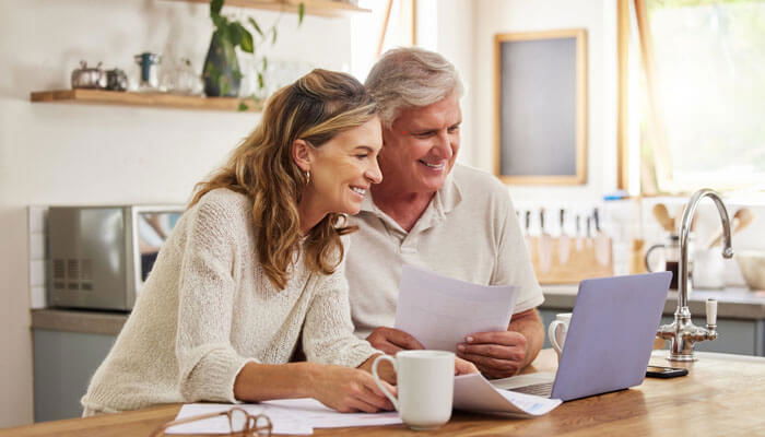 Smiling senior couple sitting at a kitchen table, reviewing documents and using a laptop, symbolizing retirement planning.