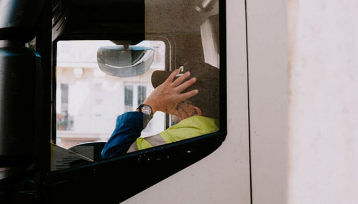 A truck driver in a high-visibility jacket, sitting inside a truck resting his hand on his head, perhaps reflecting on all the hos violations.