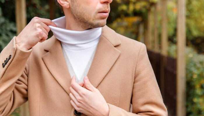 Man adjusting a beige coat over a white turtleneck sweater, showing men's sweaters in an outdoor environment.