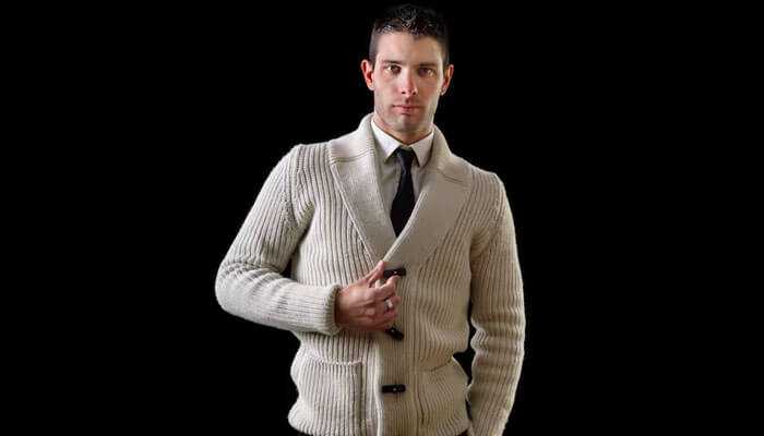 Man in cream coloured buttoned cardigan over shirt and tie, posed against black background.