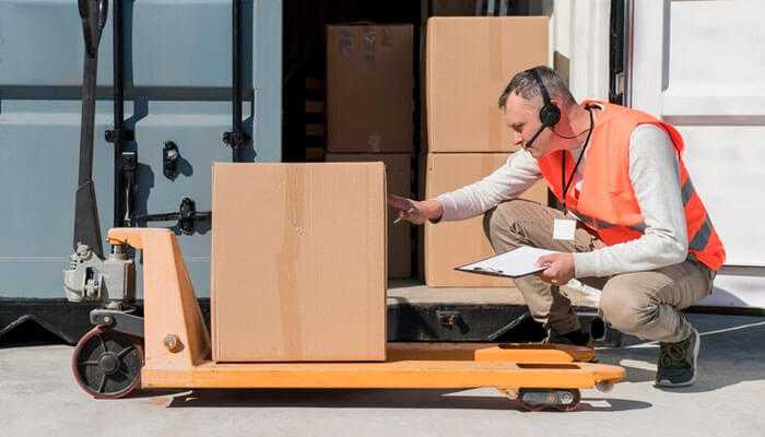 Shipping oversized goods a logistics worker wearing an orange safety vest and headset inspecting a large cardboard box on a manual pallet jack, with a clipboard in hand, standing near a truck dock