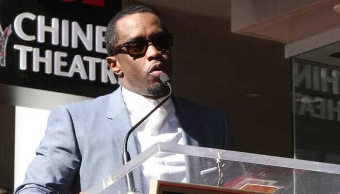 The image depicts p diddy net worth wearing sunglasses and a light-colored suit speaking at a podium with a microphone. The background shows part of a sign reading chinese theatre