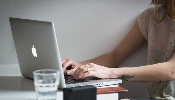 Person typing on a silver macbook at a desk with a glass of water, representing tasks common in service based businesses.