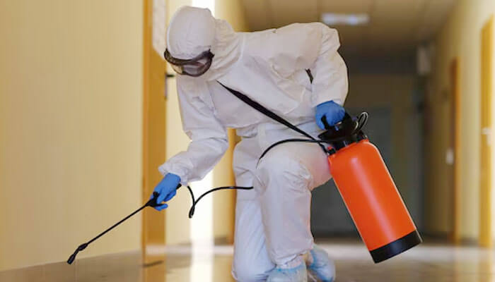 The picture depicts a person in protective gear using an orange sprayer to sanitize or decontaminate an indoor hallway, probably for mold remediation or pest control.