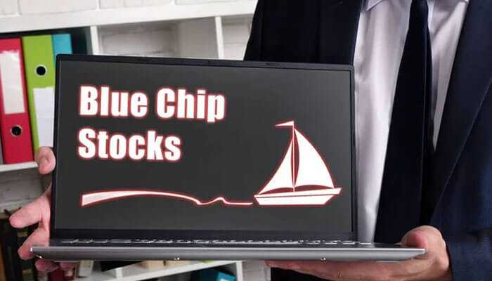 A man in suit holding a laptop with blue chip stocks and a sailboat graphic on the screen.