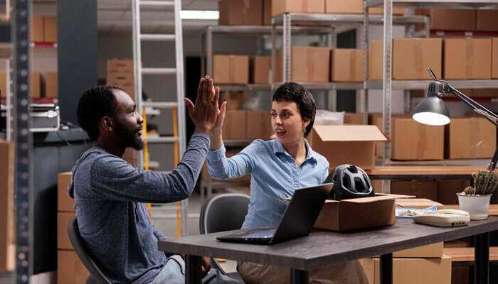 Two high-fiving coworkers in a warehouse during the supplier onboarding process, with boxes and a laptop in the background.