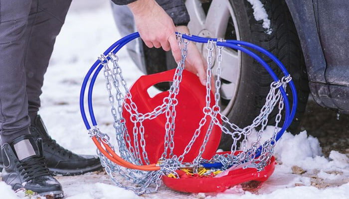 Person holding snow chains near the tire of a car, in snow, depicting following passenger vehicle chain laws.