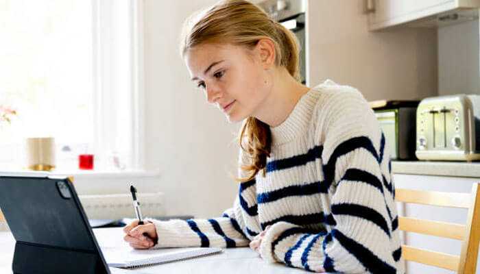 Young woman in a striped sweater studying for a levels at home, writing in a notebook while looking at a tablet on a table.