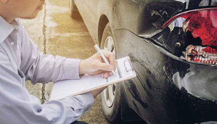 An insurance adjuster inspects a damaged car and makes notes on a clipboard, as shown in the car accident claims process.