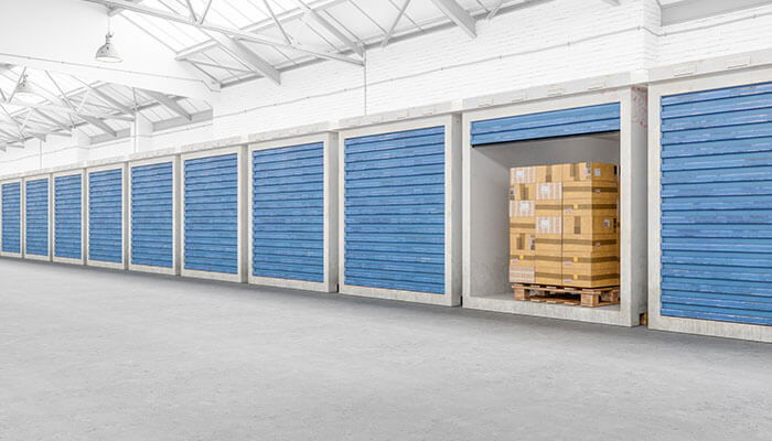 Private storage containers with blue roll-up doors, one unit open revealing stacked boxes on a pallet