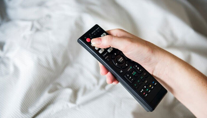 A person holding a black tv remote control, possibly for an amazon fire stick, while sitting on a bed with a white bedsheet