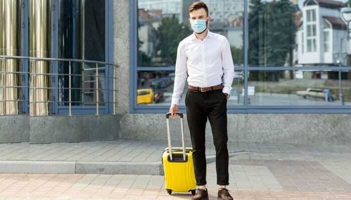 A man with a yellow suitcase and mask in an urban setting, emphasizing the need for travel insurance.