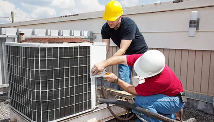 Two workers on a rooftop repair an hvac unit, demonstrating skills from hvac training programs. One wears a yellow hard hat, the other a white hard hat. It's a sunny day.