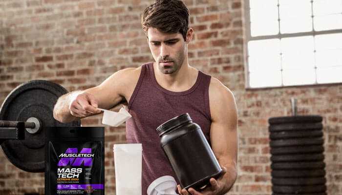 A man in the gym with a maroon tank top on, shaking a protein shake. Mass gainers muscletech mass tech extreme 2000.