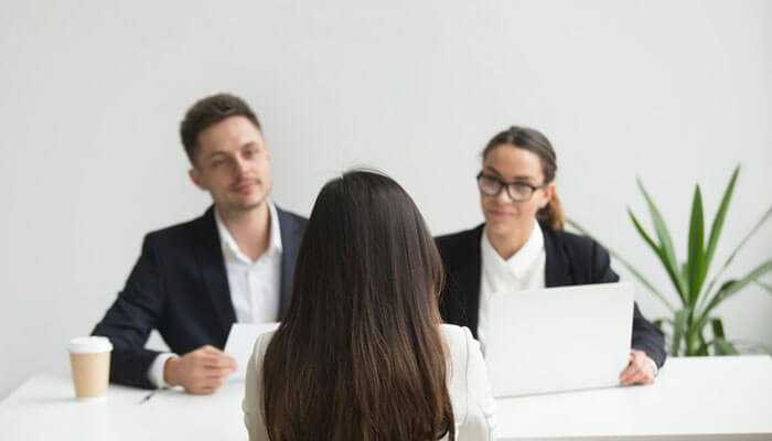 A woman sits facing two interviewers discussing common sales challenges in a professional office setting with a plant.