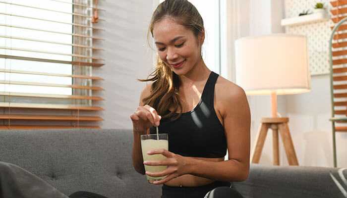 Young woman in activewear stirring a glass of collagen supplements drink while sitting on a couch, promoting bone health