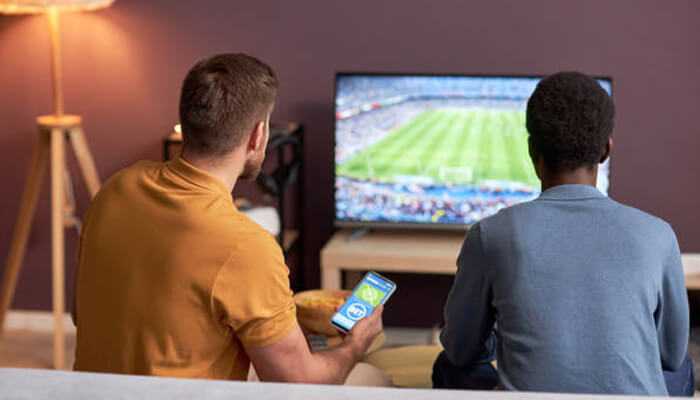 Two men watching a live football match on tv while one checks a sports betting app on his smartphone, illustrating the rise of live streaming and in-play betting in india’s sports betting industry