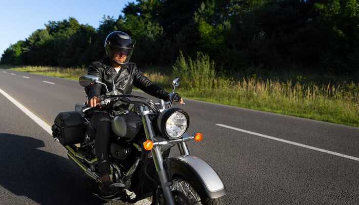 Motorcyclist wearing a helmet and leather jacket riding a motorcycle on a clear road surrounded by green trees, representing the experience of holding a wyoming motorcycle license.
