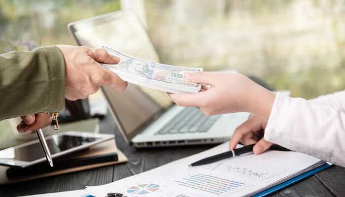 Close-up of hands exchanging cash over financial charts and a laptop, representing real-world access to pedirayudas government financial support initiatives.