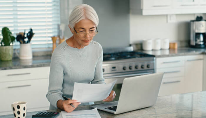 An older woman reviewing paperwork related to her life insurance policy while working on a laptop in the kitchen.