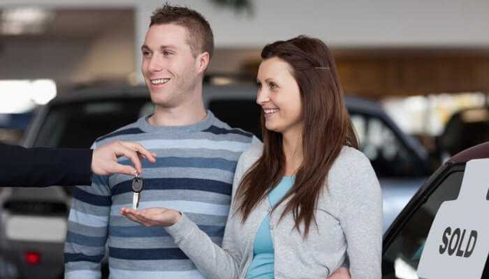 A happy couple receiving car keys after buying a second hand vehicle, with a'SOLD' sign visible in the background.