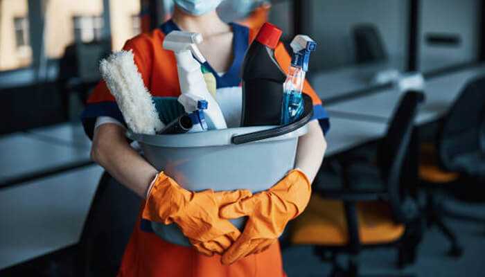 A person wearing protective gloves, holding a bucket filled with commercial cleaning products like sprays, brushes, and cleaning solutions in an office setting.