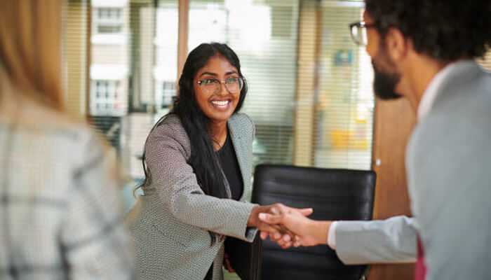 Executive assistant greeting a new colleague with a handshake in a corporate office setting.