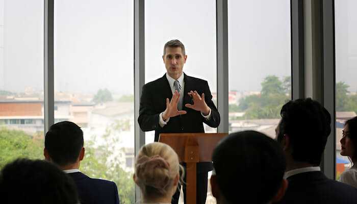 Engaging motivational speaker in a business suit speaking energetically to an audience in a bright, nature-filled indoor setting.