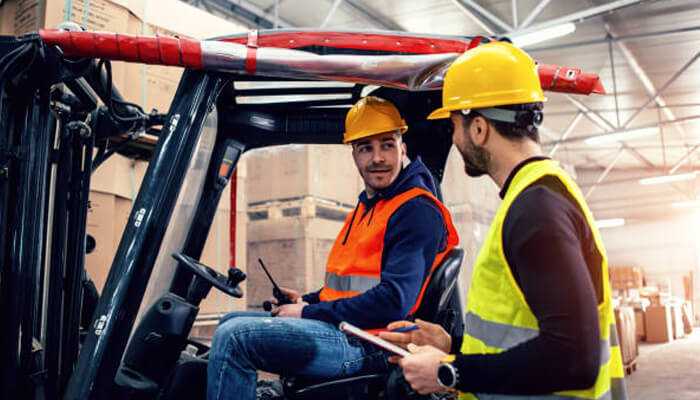 Two warehouse workers discussing procedures near a forklift, emphasizing communication and awareness of safety risks for warehouses.