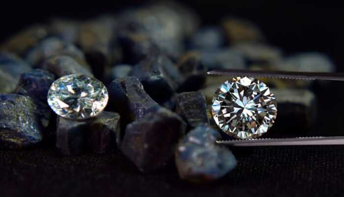 Two polished diamonds, one held by tweezers and the other resting on uncut stones, showcasing brilliance and clarity—explore more about the diamond cut grades that determine their sparkle and value.